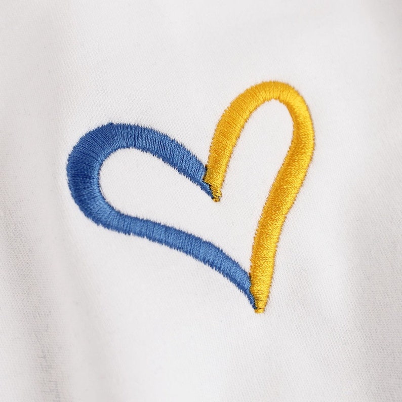 May include: A white fabric background with a blue and yellow embroidered heart.
