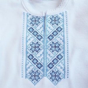 Puede incluir: Camisa blanca bordada con un patr&oacute;n floral azul y blanco. El bordado es de estilo ucraniano tradicional.