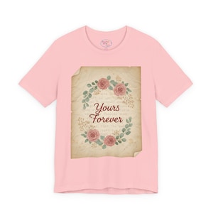Może przedstawiać: Jasnoróżowa koszulka z wzorem w stylu vintage. Wzór zawiera wieniec kwiatowy z różowymi różami i słowami "Yours Forever" na tle przypominającym pergamin. Koszulka ma krótkie rękawy i okrągły dekolt.