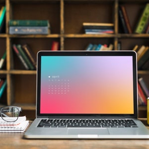 Puede incluir: Un ordenador portátil plateado con un calendario en la pantalla. La pantalla muestra un fondo degradado con un calendario para abril. El portátil está sobre una mesa de madera con una taza amarilla y una pila de cuadernos.