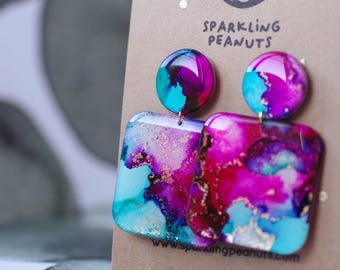 May include: A pair of colorful earrings with a square and round design. The earrings feature a vibrant mix of turquoise, magenta, and gold, with the text "Sparkling Peanuts" on the backing card. The earrings are a unique accessory.