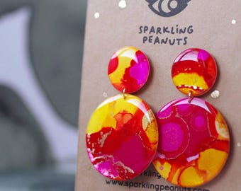 May include: A pair of round drop earrings featuring a vibrant abstract pattern in shades of hot pink, bright yellow, and orange. The earrings are displayed on a brown card printed with the text Sparkling Peanuts and an eye icon.