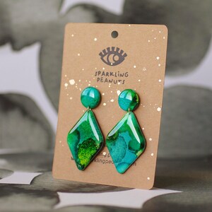 May include: A pair of green and teal diamond-shaped earrings with a marbled effect, displayed on a brown card with the text "Sparkling Peanuts". The earrings have a round stud and a geometric drop.