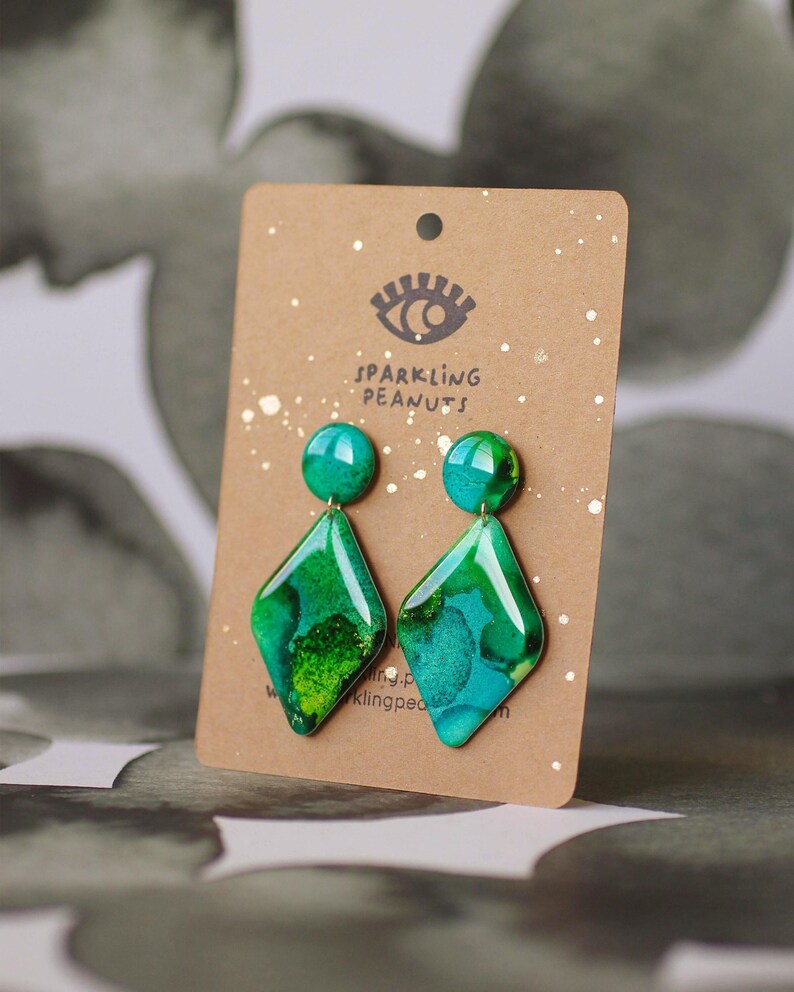May include: A pair of green and teal diamond-shaped earrings with a marbled effect, displayed on a brown card with the text "Sparkling Peanuts". The earrings have a round stud and a geometric drop.