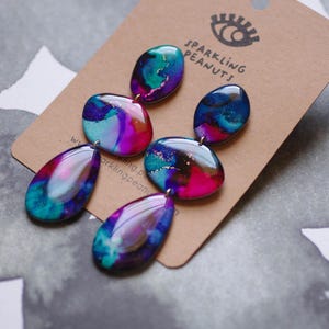 May include: Abstract design earrings in teal, purple, and pink. The earrings have a glossy finish and are attached to a brown card. The card features the text "Sparkling Peanuts".