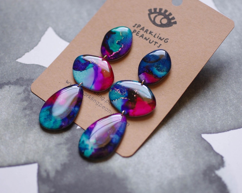 May include: Abstract design earrings in teal, purple, and pink. The earrings have a glossy finish and are attached to a brown card. The card features the text "Sparkling Peanuts".