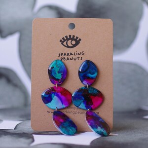 May include: Abstract earrings in blue, purple, and pink, with a marbled design. The earrings are displayed on a brown card with the text "Sparkling Peanuts" and a website address. The earrings are a unique accessory.