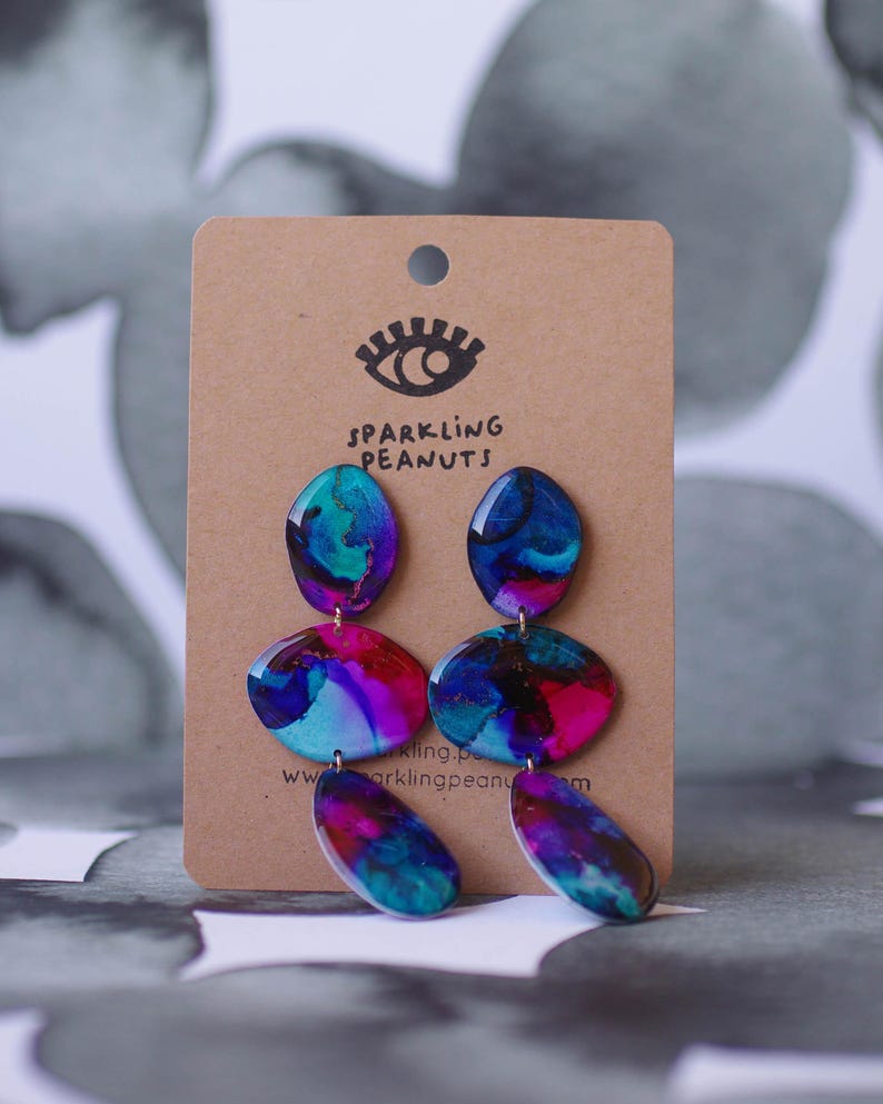 May include: Abstract earrings in blue, purple, and pink, with a marbled design. The earrings are displayed on a brown card with the text "Sparkling Peanuts" and a website address. The earrings are a unique accessory.