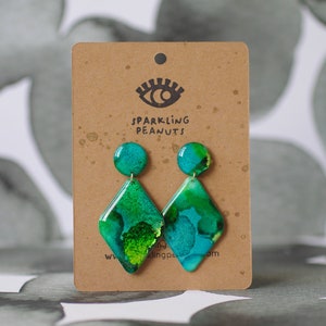 May include: A pair of handmade earrings with a teal and green abstract design. The earrings have a round stud and a diamond-shaped pendant. The earrings are displayed on a brown card with the text "Sparkling Peanuts".