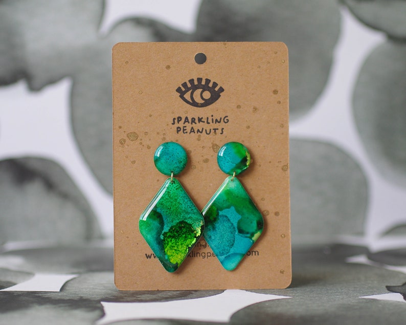 May include: A pair of handmade earrings with a teal and green abstract design. The earrings have a round stud and a diamond-shaped pendant. The earrings are displayed on a brown card with the text "Sparkling Peanuts".