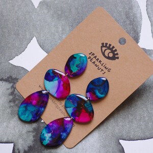 May include: Abstract design earrings in teal, purple, and pink. The earrings are attached to a brown card with the text "SPARKLING PEANUTS" and a logo. The earrings have a glossy finish. The earrings are a unique accessory.