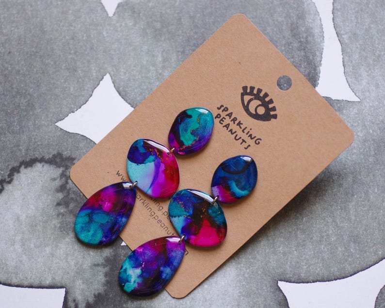 May include: Abstract design earrings in teal, purple, and pink. The earrings are attached to a brown card with the text "SPARKLING PEANUTS" and a logo. The earrings have a glossy finish. The earrings are a unique accessory.