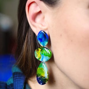 May include: Dangle earrings featuring three stones with an abstract design. The top stone is blue, the middle is green and yellow, and the bottom is green and blue. The earrings are worn on a person's ear.