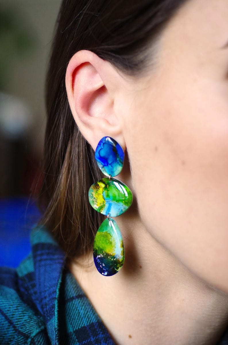 May include: Dangle earrings featuring three stones with an abstract design. The top stone is blue, the middle is green and yellow, and the bottom is green and blue. The earrings are worn on a person's ear.