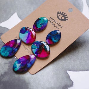 May include: Dangle earrings with a marbled design in teal, purple, and pink. The earrings are attached to a brown tag with the text "Sparkling Peanuts" and an eye logo.