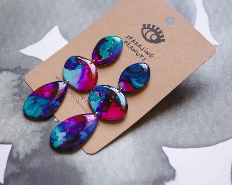 May include: Dangle earrings with a marbled design in teal, purple, and pink. The earrings are attached to a brown tag with the text "Sparkling Peanuts" and an eye logo.
