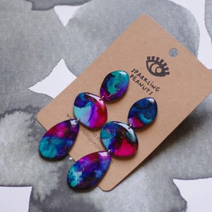 May include: Abstract earrings in blue, teal, pink, and purple with a marbled effect. The earrings are displayed on a brown card with the text "Sparkling Peanuts" and an eye logo.