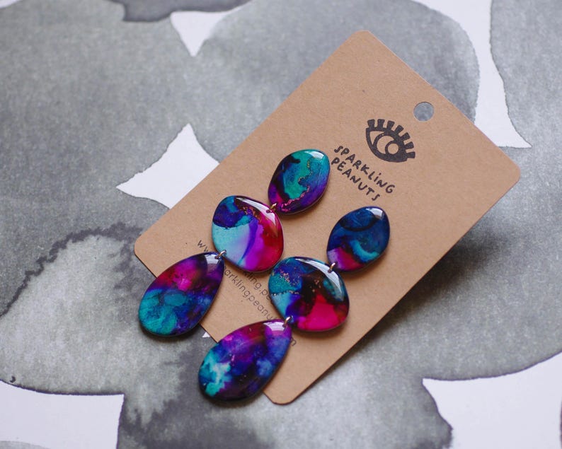 May include: Abstract earrings in blue, teal, pink, and purple with a marbled effect. The earrings are displayed on a brown card with the text "Sparkling Peanuts" and an eye logo.