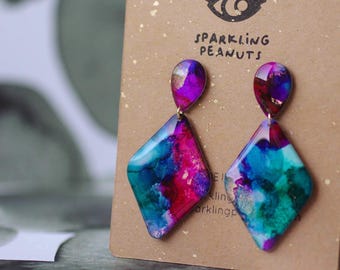 May include: Colorful abstract earrings with a teardrop top and a diamond-shaped bottom. The earrings feature vibrant hues of blue, pink, purple, and gold. The earrings are displayed on a brown card with the text "Sparkling Peanuts".
