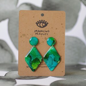 May include: A pair of handmade earrings with a teal and green abstract design. The earrings are diamond-shaped with a circular top, attached to a brown card with the text "SPARKLING PEANUTS" and an eye logo.