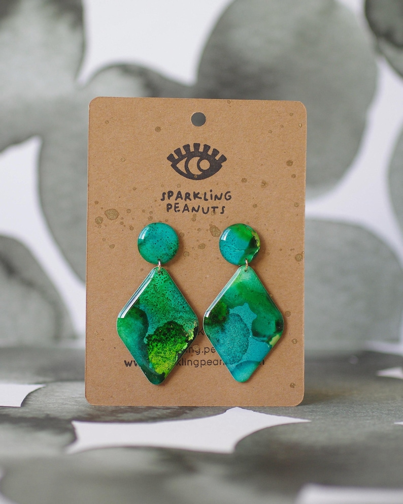 May include: A pair of handmade earrings with a teal and green abstract design. The earrings are diamond-shaped with a circular top, attached to a brown card with the text "SPARKLING PEANUTS" and an eye logo.