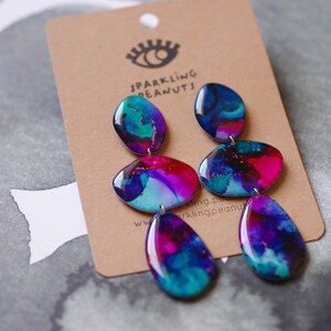 May include: Abstract design earrings in shades of blue, teal, pink, and purple. The earrings have a series of connected, glossy, oval-shaped pendants. They are attached to a brown card with the text "Sparkling Peanuts" and a logo of an eye.