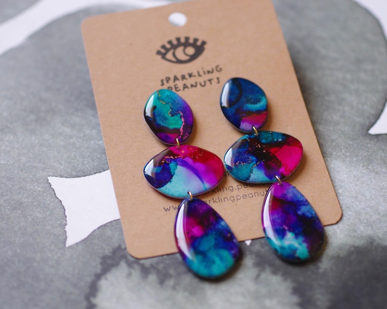 May include: Abstract design earrings in shades of blue, teal, pink, and purple. The earrings have a series of connected, glossy, oval-shaped pendants. They are attached to a brown card with the text "Sparkling Peanuts" and a logo of an eye.