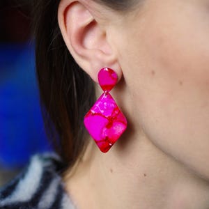 May include: Hot pink and red geometric earrings. The earrings feature a small, rounded stud and a larger, diamond-shaped pendant. The earrings are made of a translucent material with a marbled effect. The earrings are shown on a person.