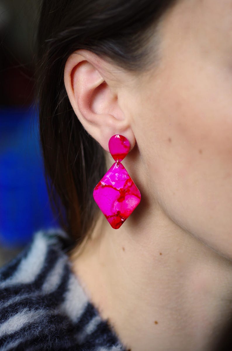 May include: Hot pink and red geometric earrings. The earrings feature a small, rounded stud and a larger, diamond-shaped pendant. The earrings are made of a translucent material with a marbled effect. The earrings are shown on a person.