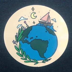 Könnte beinhalten: Ein beiger Aufkleber mit einem blauen und grünen Globus, umgeben von grünen Blättern. Ein rosa und braunes Segelboot segelt auf dem Globus. Der Hintergrund ist hellbeige mit einer Mondsichel und Sternen.