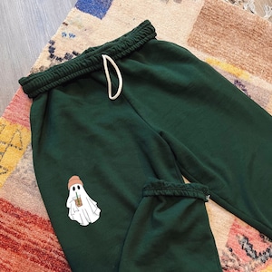 May include: Dark green sweatpants with a white ghost graphic wearing a pink hat and holding a brown beverage.