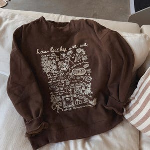 May include: A brown sweatshirt with the text "how lucky are we" and a white graphic design of flowers, leaves, and other illustrations. The long-sleeved top is made of a soft material and is folded on a white couch.