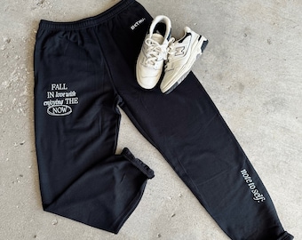 May include: Black sweatpants with white text that reads "Fall in love with enjoying the now" on the left leg and "note to self" on the right leg. A pair of white sneakers are lying on top of the sweatpants.