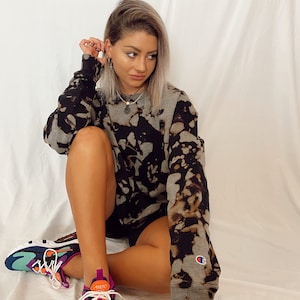 Puede incluir: Una mujer con una sudadera gris y negra con estampado tie-dye, un logotipo de Champion en la manga, pantalones cortos negros y zapatillas Nike multicolores.