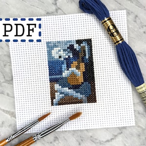 May include: A white Aida cloth with a cross-stitch pattern of a blue and brown abstract design. The cloth is surrounded by two paintbrushes and a spool of blue embroidery floss.