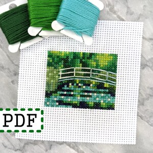 Puede incluir: Un tejido blanco Aida con un patrón de punto de cruz de un puente verde sobre un estanque con nenúfares. Se incluyen tres carretes de hilo para bordar: verde, verde oscuro y turquesa.