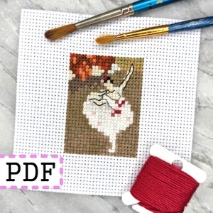 May include: A cross-stitch pattern featuring a ballerina in a white tutu with pink accents, standing on a brown stage with a red background. The pattern is printed on white Aida cloth and includes a spool of red embroidery floss.