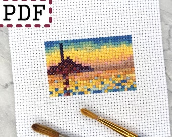 May include: A white Aida cloth with a colorful cross-stitch pattern of a sunset landscape. Two paint brushes are lying on the cloth.