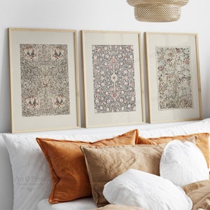 以下が含まれることがあります： グレー、ブラウン、ピンクの複雑な花柄が描かれた額装されたプリントが3枚。プリントは、白の壁に、ブラウンとホワイトの枕のあるベッドの上に飾られています。
