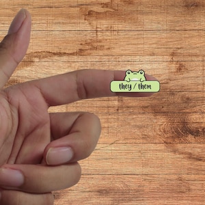 May include: A green frog wearing a light green banner with the text "they/them" on a wooden background.