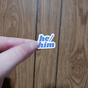 Puede incluir: Una pegatina blanca con texto azul que dice "he/him" en una fuente retro.