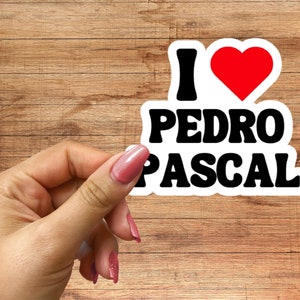 May include: White sticker with black text that reads "I love Pedro Pascal" with a red heart in between the words "I" and "love".