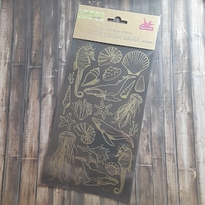 Puede incluir: Un paquete de transferencias metálicas para frotar con ilustraciones de criaturas marinas. Los diseños incluyen caballitos de mar, ballenas, medusas, estrellas de mar y conchas. El paquete tiene el texto "Crafts Square" y "rub-on transfers".
