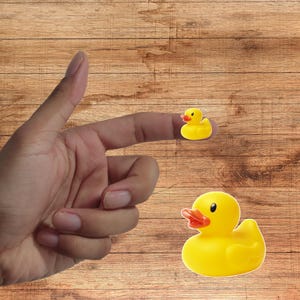 Puede incluir: Dos patos de goma amarillos con picos naranjas y ojos negros. Uno está en un dedo. Los patos están sobre una superficie de madera.