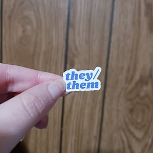 Puede incluir: Una pequeña pegatina con las palabras "they/them" en azul. El texto está en una fuente redondeada sin serifa, con un borde blanco. La pegatina se sostiene en una mano sobre un fondo de madera.