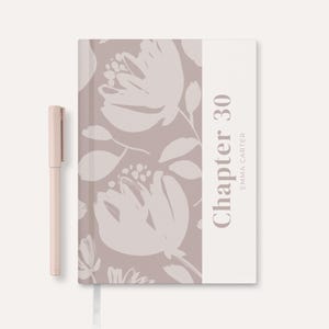 May include: A blush-colored pen rests beside a floral-patterned journal. The journal cover features a large, light-colored floral design on a mauve background. The text "Chapter 30" and "Emma Carter" are printed vertically on a white stripe.