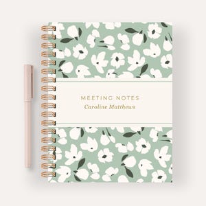 May include: A floral patterned notebook with a gold spiral binding and a matching pen. The notebook features a light green background with white and dark green floral designs. The text "MEETING NOTES Caroline Matthews" is displayed on a cream-colored band.