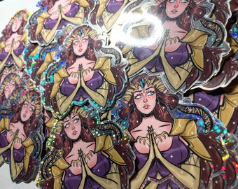 May include: A stack of holographic stickers featuring a woman with long brown hair, wearing a purple dress and gold armor. She has her hands clasped in front of her, and her eyes are closed. The stickers have a glittery finish.