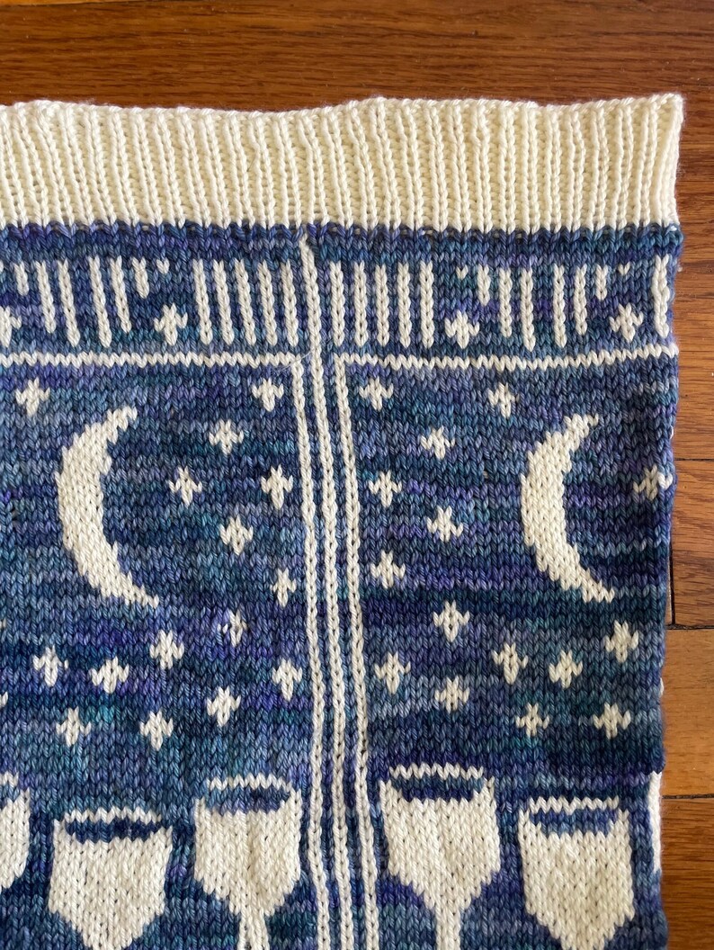 Pu&ograve; includere: Una coperta di maglia con un motivo blu e bianco con lune, stelle e tazze. La coperta ha un bordo bianco.