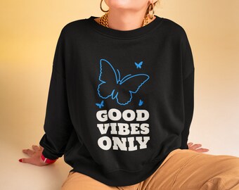 May include: Black sweatshirt with a blue butterfly graphic and the words "GOOD VIBES ONLY" in white. The sweatshirt is paired with tan pants. The person is wearing orange sunglasses and gold jewelry.